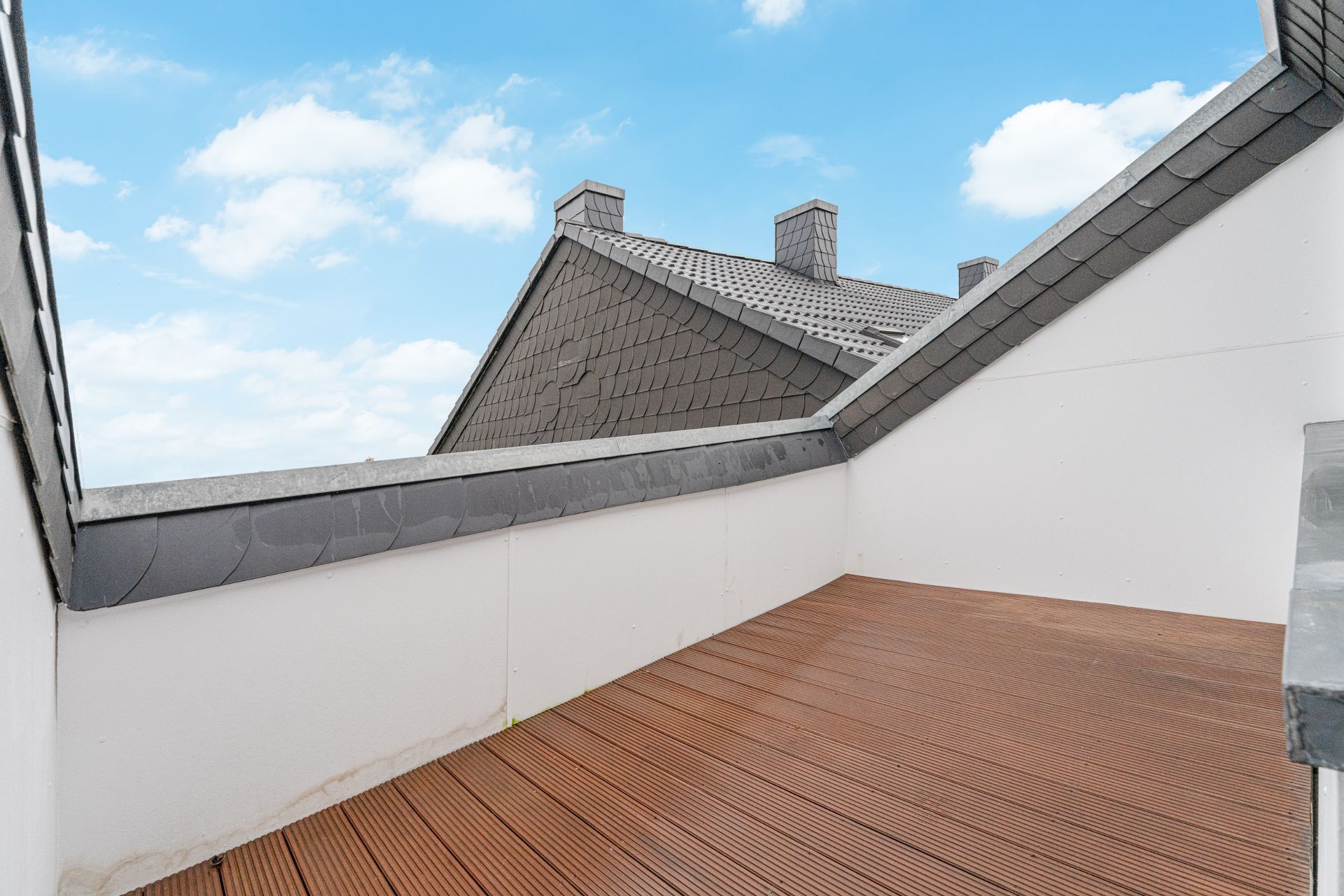 Kleine Dachterrasse mit Holzboden und Blick auf Nachbardächer unter blauem Himmel.