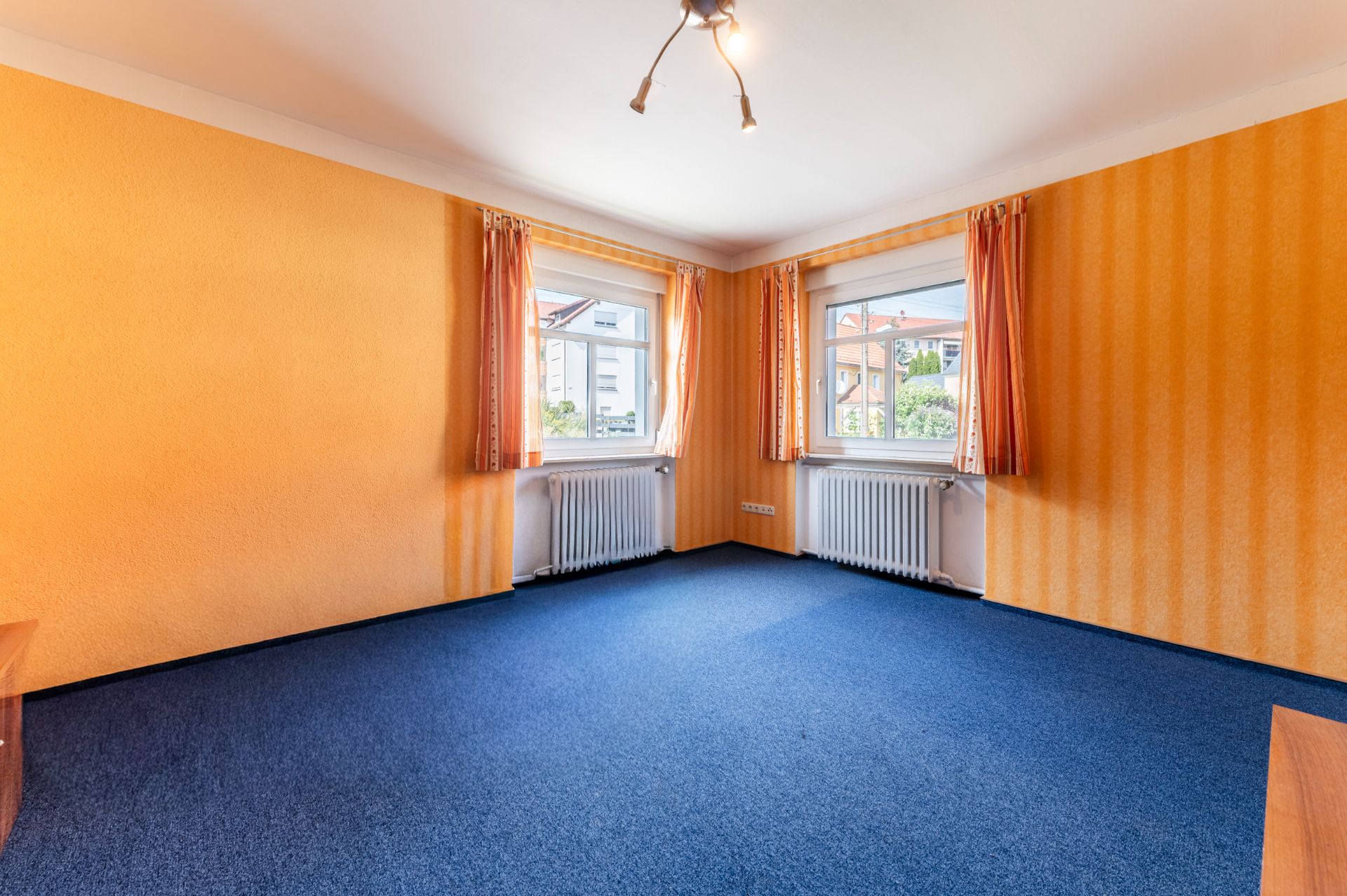 Leeres Zimmer mit orangefarbenen Wänden, zwei Fenstern mit Vorhängen und blauem Teppichboden.