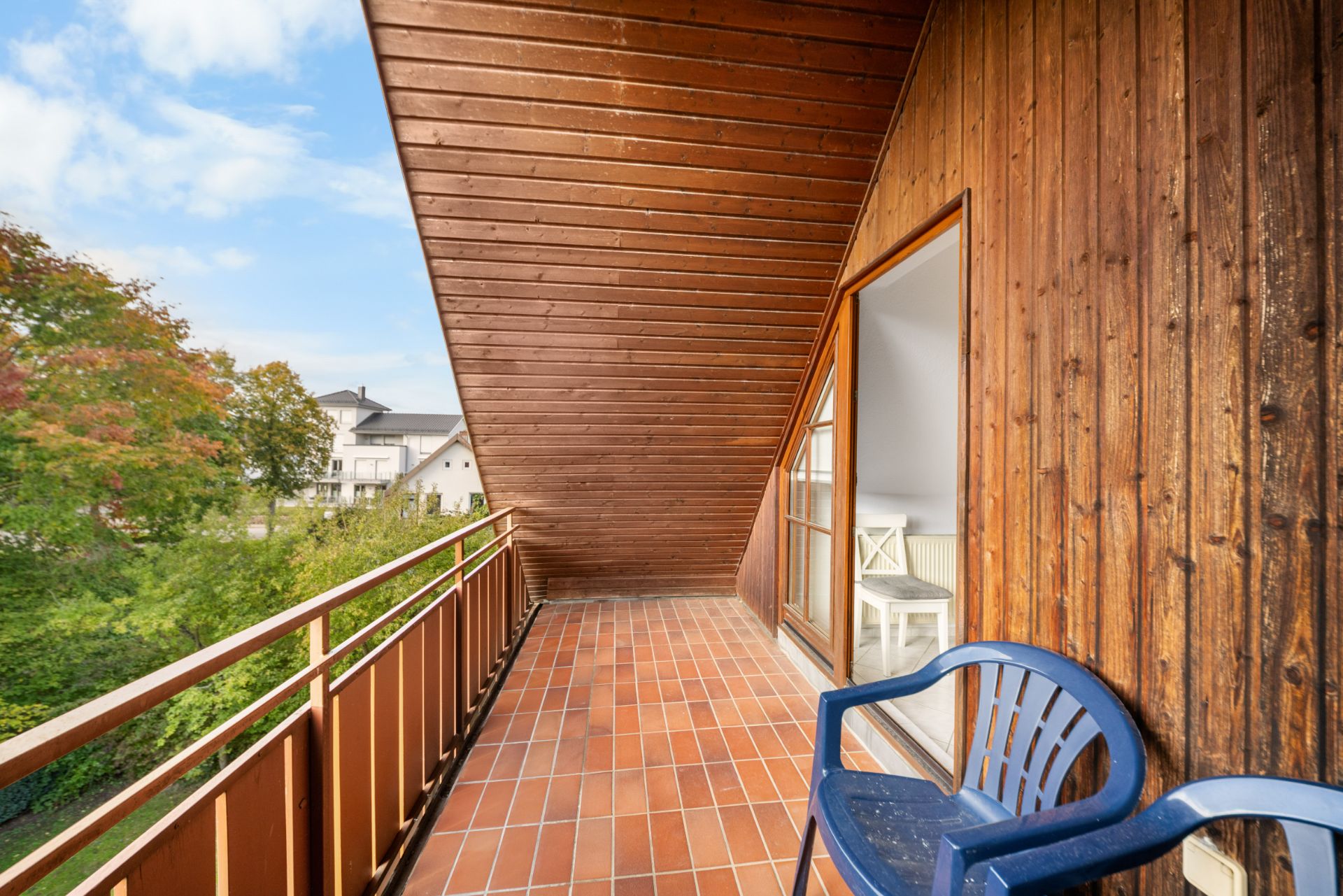 Überdachter Balkon mit Holzwand, Terrakottafliesen und einem blauen Plastikstuhl.