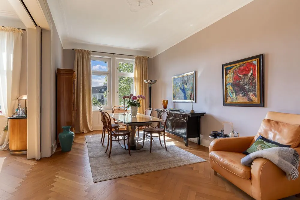 Esszimmer mit Holztisch, Stühlen, braunem Sessel und großen Fenstern, dekoriert mit Gemälden und Blumen.
