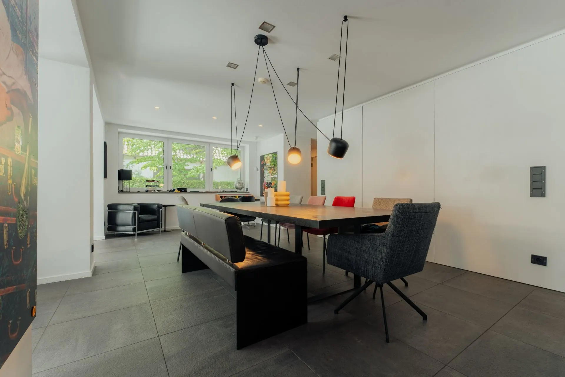 Modernes Esszimmer mit großem Tisch, verschiedenen Stühlen, Hängelampen und Fenster im Hintergrund.