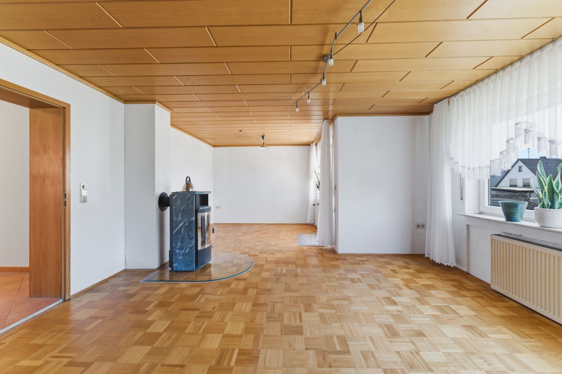 Helles Wohnzimmer mit Holzdecke, Parkettboden, Kamin und großem Fenster mit Vorhang und Pflanzen.