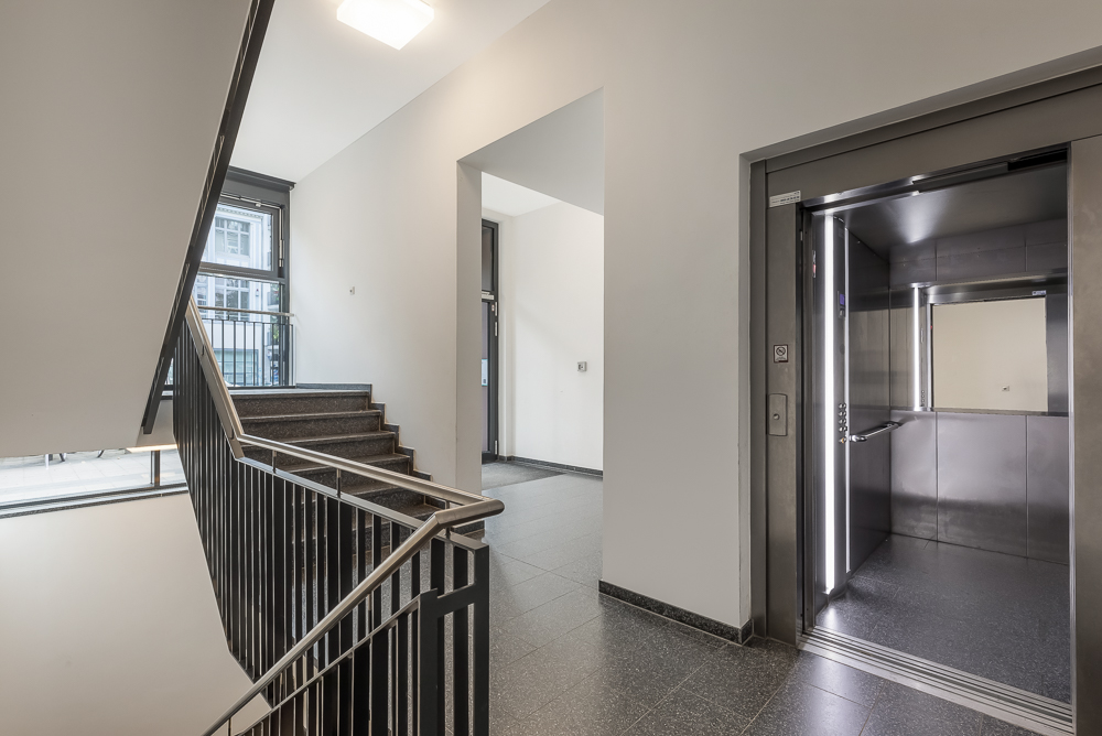 Heller Hausflur mit Treppenaufgang und offenem Aufzug, Fenster im Hintergrund.