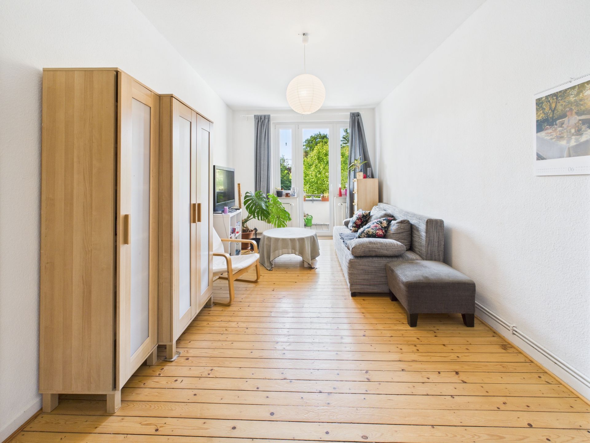 Helles Wohnzimmer mit Holzboden, Sofa, Schrank, Sessel, Tisch und großem Fenster mit Blick ins Grüne.