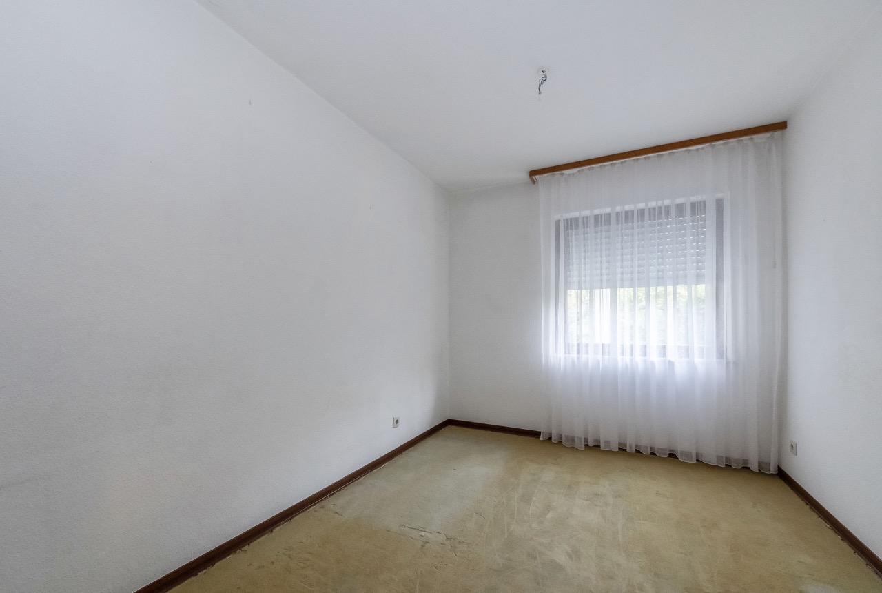 Leeres Zimmer mit hellem Teppichboden, weißer Wand und Fenster mit Vorhang.