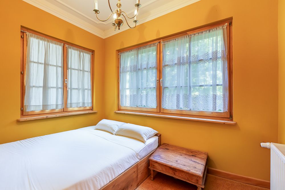 Kleines Schlafzimmer mit gelben Wänden, Bett, zwei Fenstern mit Gardinen und Holztisch.