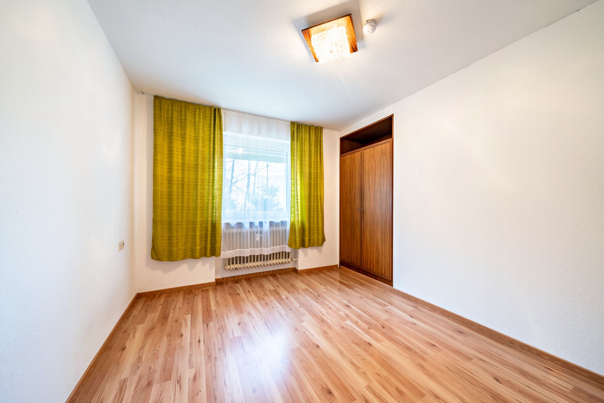 Leeres Zimmer mit Holzfußboden, großem Fenster mit grünen Vorhängen und Einbauschrank.