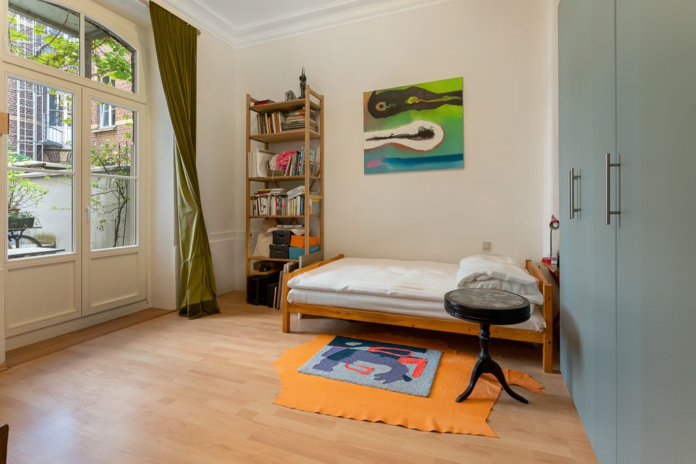 Helles Schlafzimmer mit Holzboden, Einzelbett, Regal, großem Fenster und buntem Teppich.