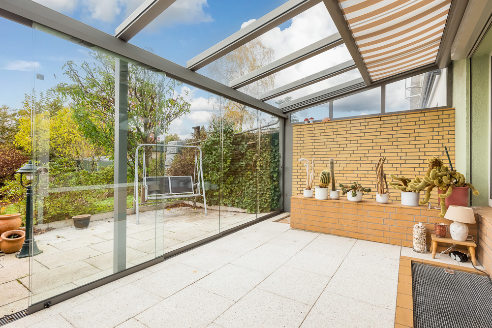 Überdachte Terrasse mit Glaswänden, Sitzbank, Kakteentöpfen und Blick auf Garten.