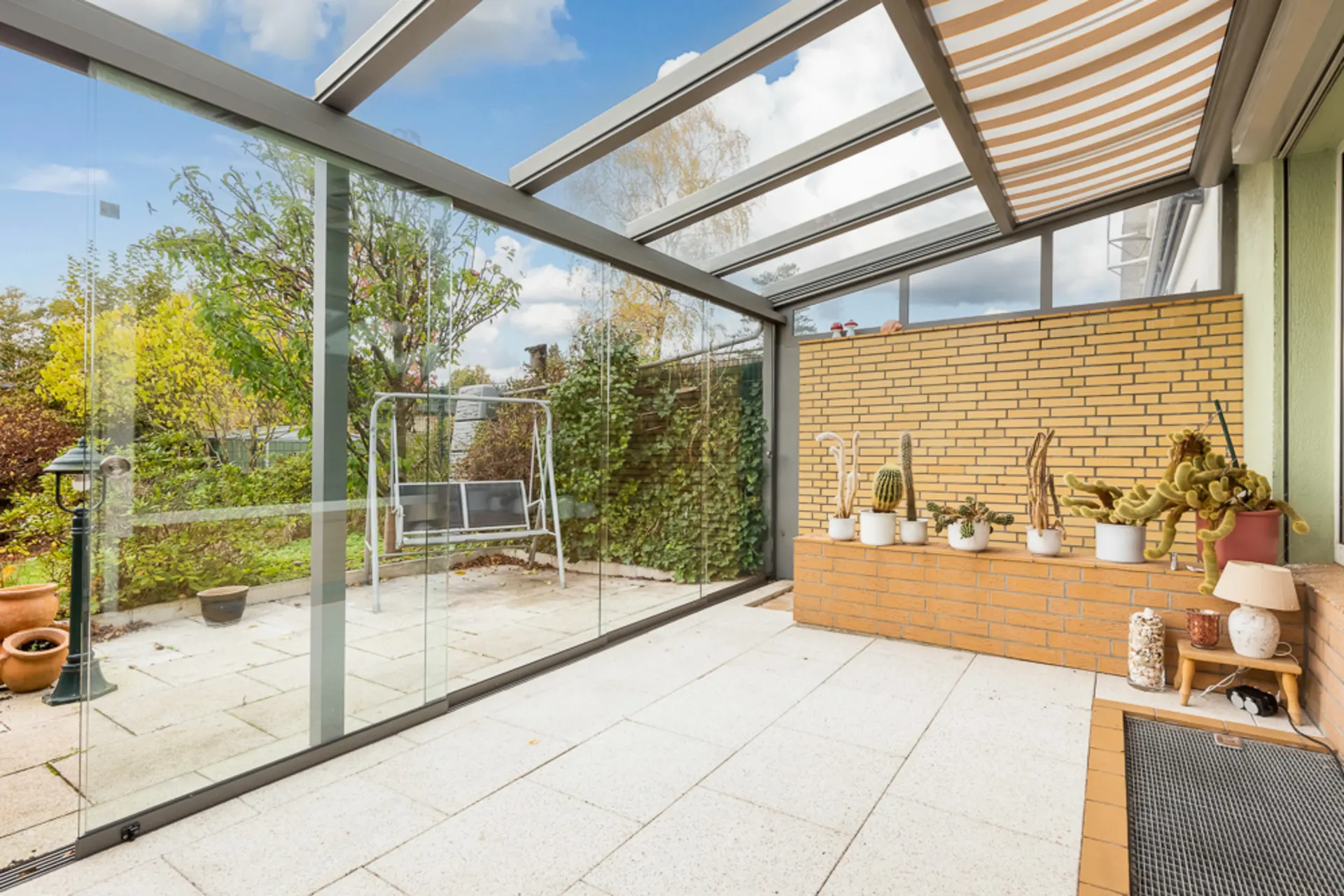 Überdachte Terrasse mit Glaswänden, Sitzbank, Kakteentöpfen und Blick auf Garten.