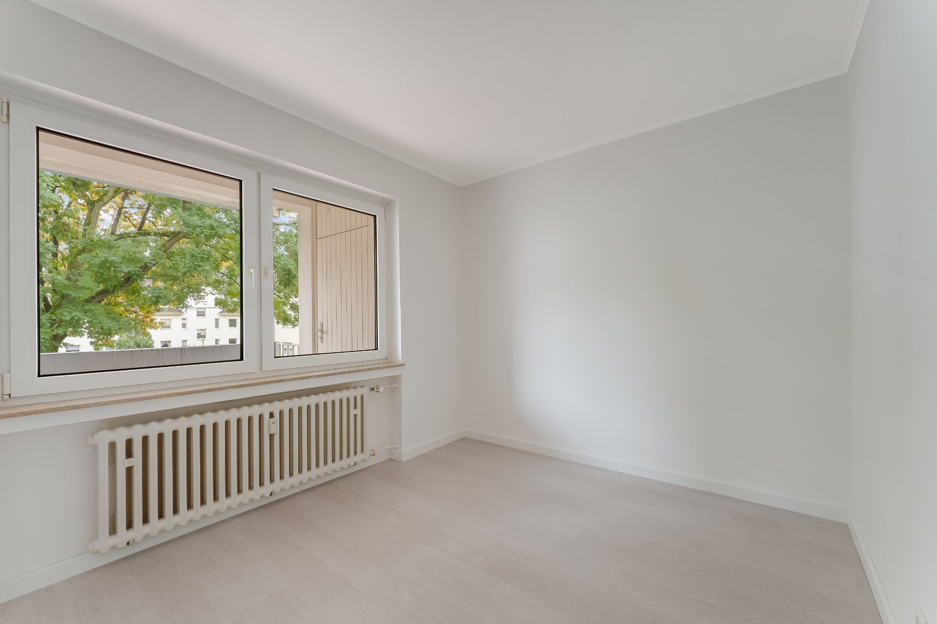 Helles, leeres Zimmer mit großem Fenster, weißem Heizkörper und hellem Boden.