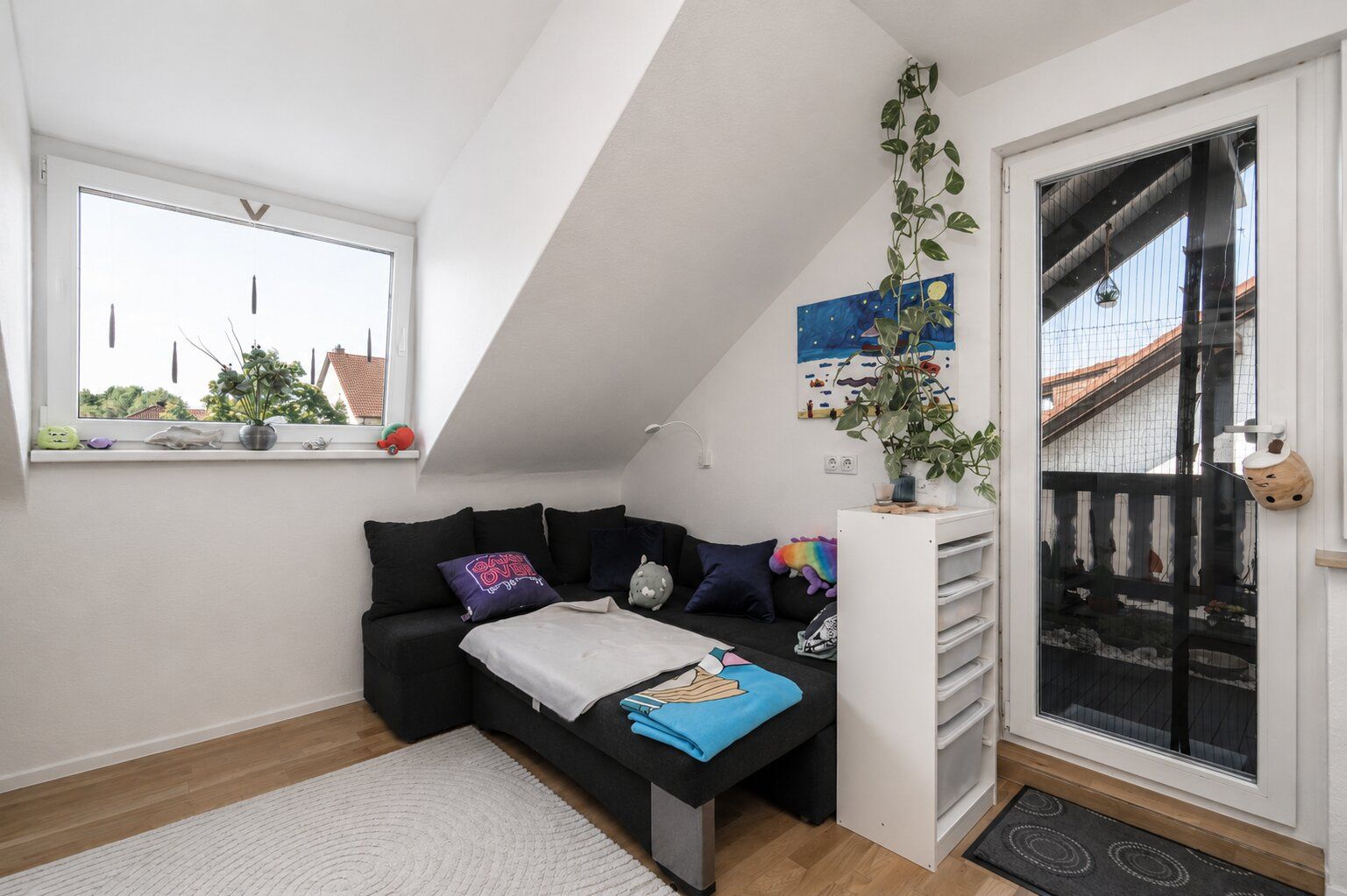 Kleines Wohnzimmer mit Ecksofa, Pflanze, Fenster und Tür zum Balkon unter Dachschräge.