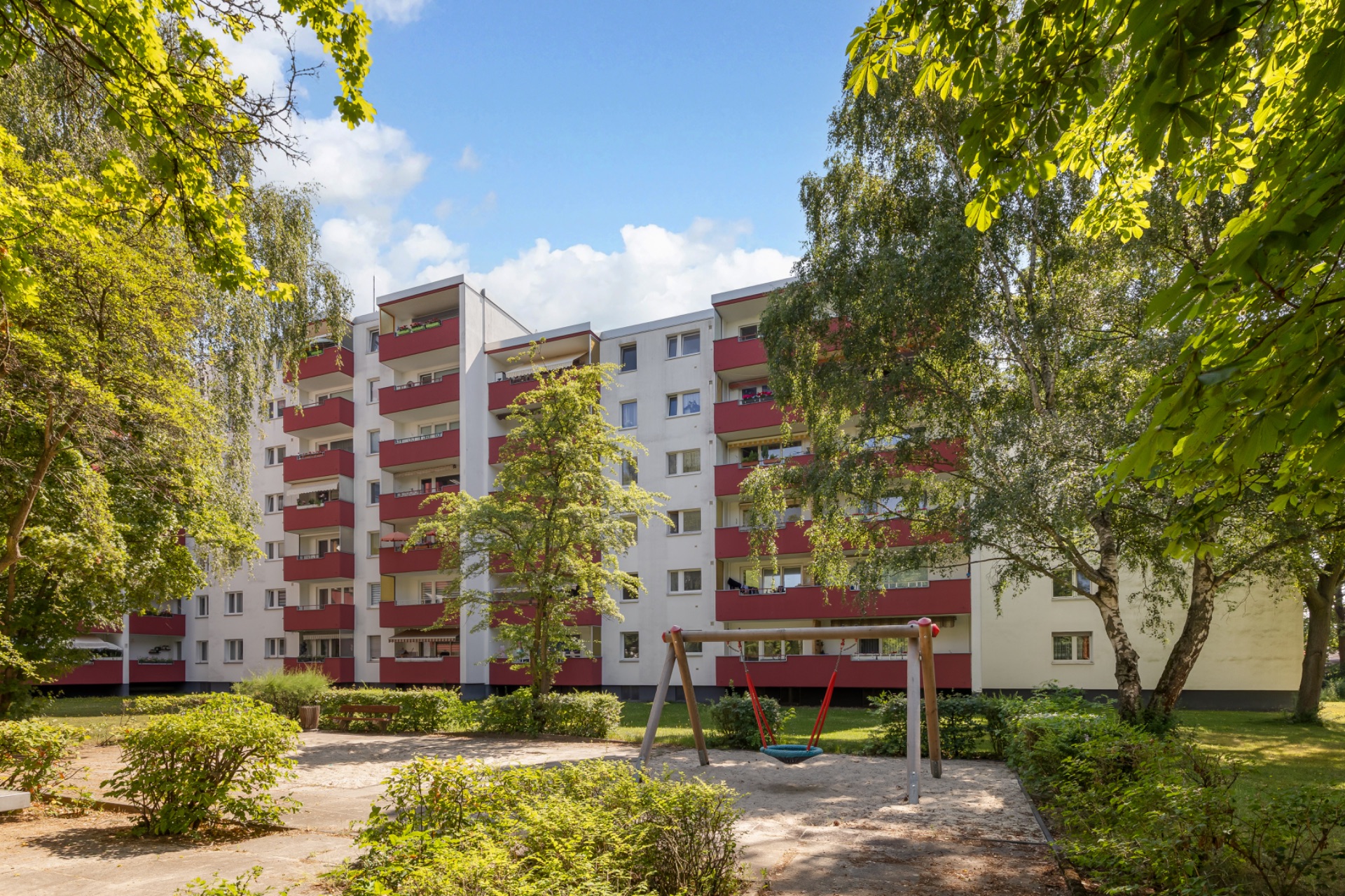 Mehrfamilienhaus mit roten Balkonen und Schaukel im grünen Gartenbereich.