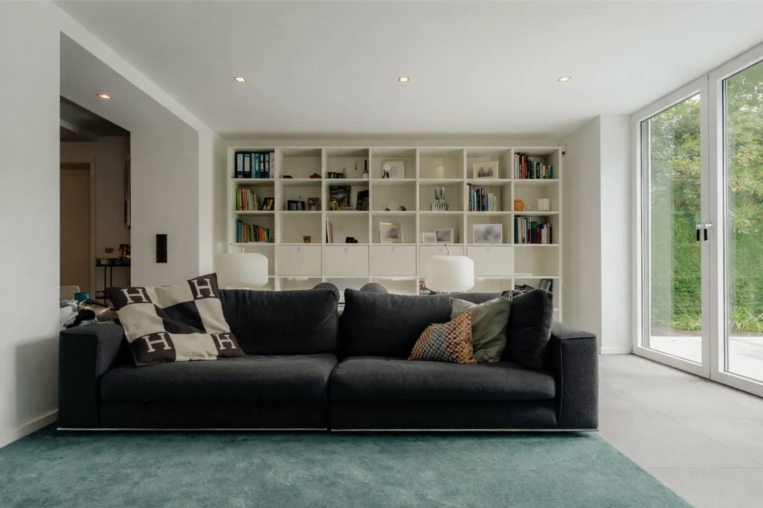 Wohnzimmer mit dunkelgrauem Sofa, Kissen, Regalwand und viel Tageslicht durch große Fenster.