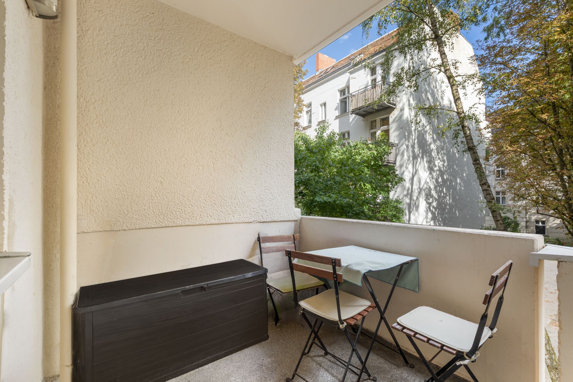 Balkon mit zwei Stühlen, kleinem Tisch und einer schwarzen Aufbewahrungsbox, Blick auf Bäume und Nachbarhaus.