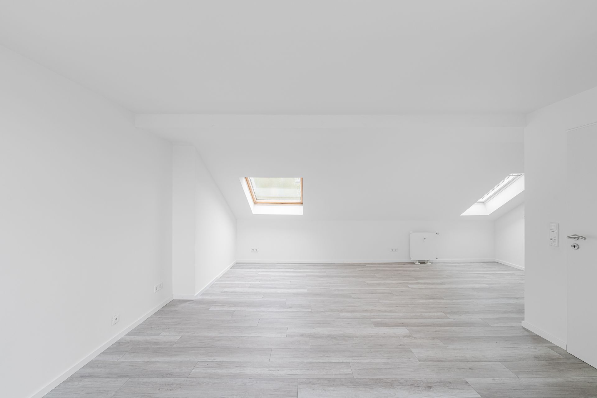 Heller leerer Raum mit schrägem Dach, Dachfenstern und hellem Holzboden.