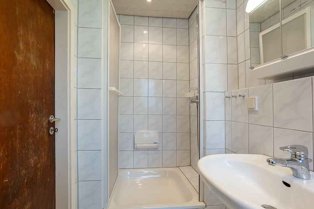 Kleines Bad mit Dusche, Waschbecken und Spiegelschrank, weiße Fliesen an den Wänden.