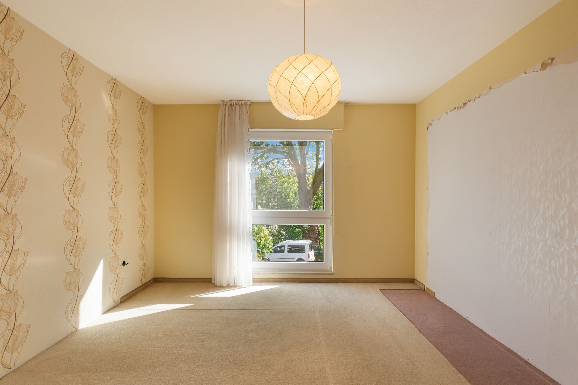 Leerer Raum mit hellem Teppich, Fenster, Vorhang und Lampe, eine Wand teils beschädigt.