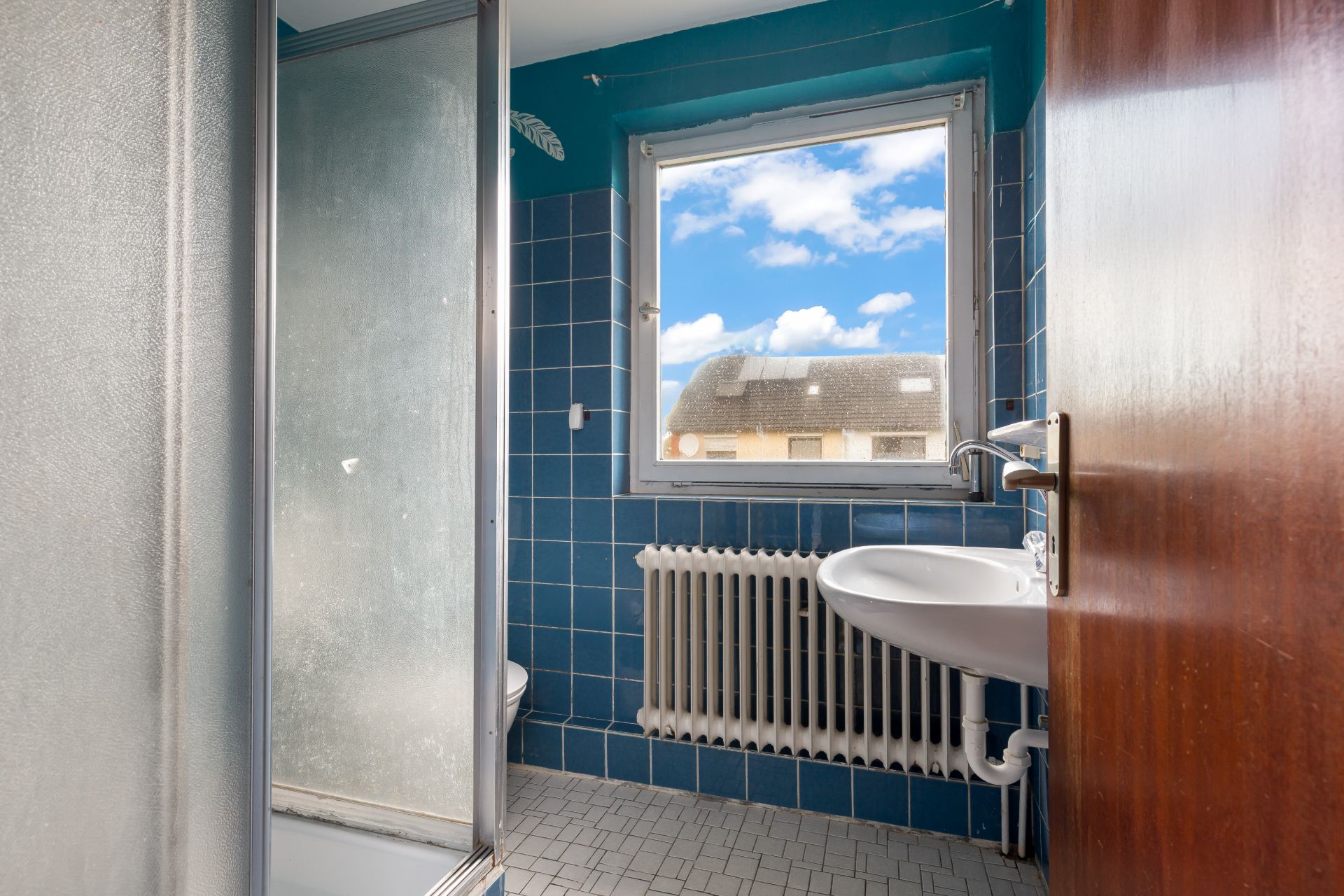 Kleines Badezimmer mit blauen Fliesen, Fenster, Waschbecken, Dusche und Heizkörper.
