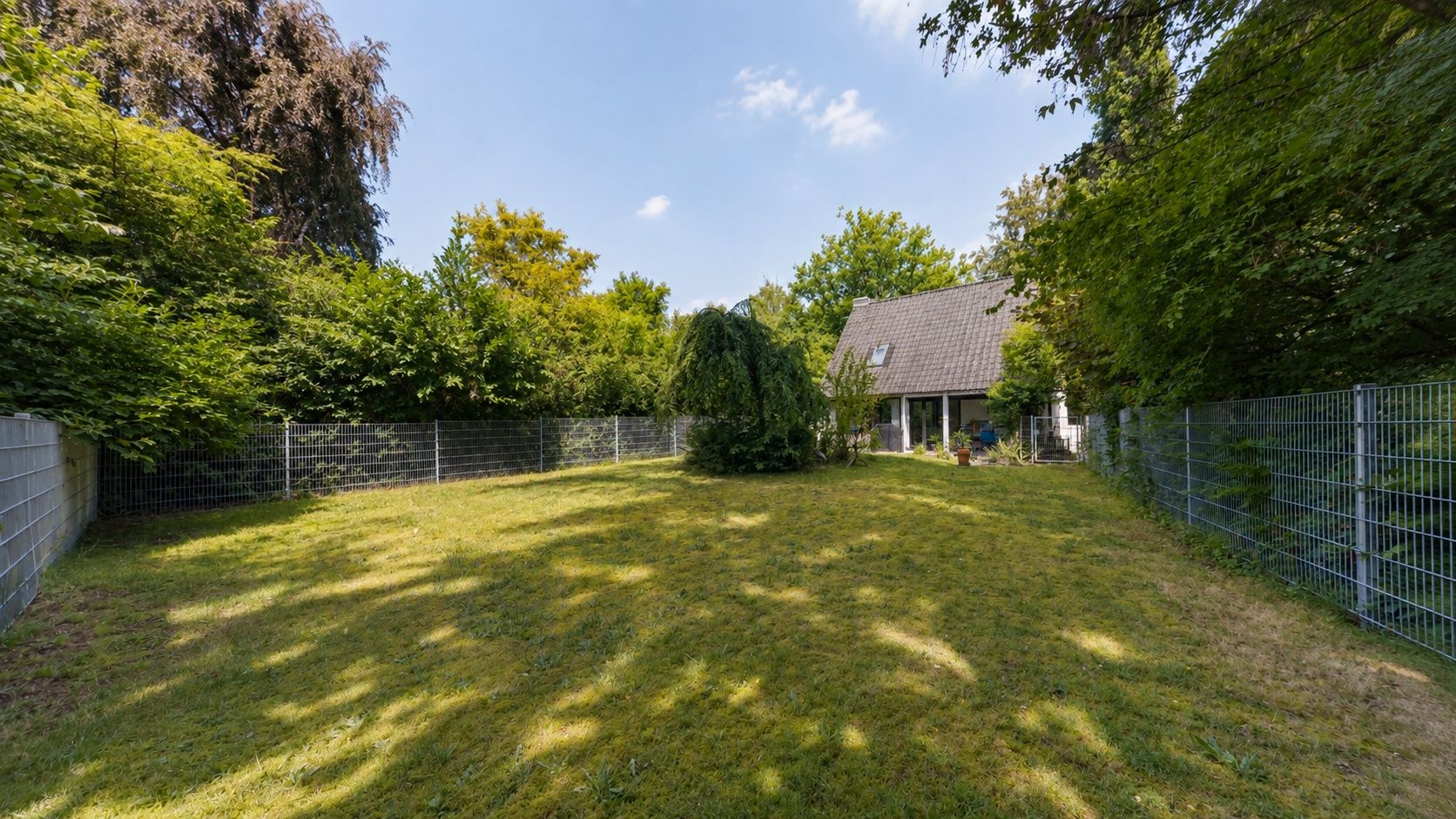 Großer, umzäunter Garten mit Rasenfläche und Haus im Hintergrund, umgeben von Bäumen.