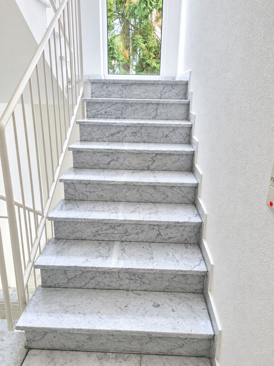 Helle Marmortreppe mit Geländer, oben ein Fenster mit Blick auf grüne Bäume.