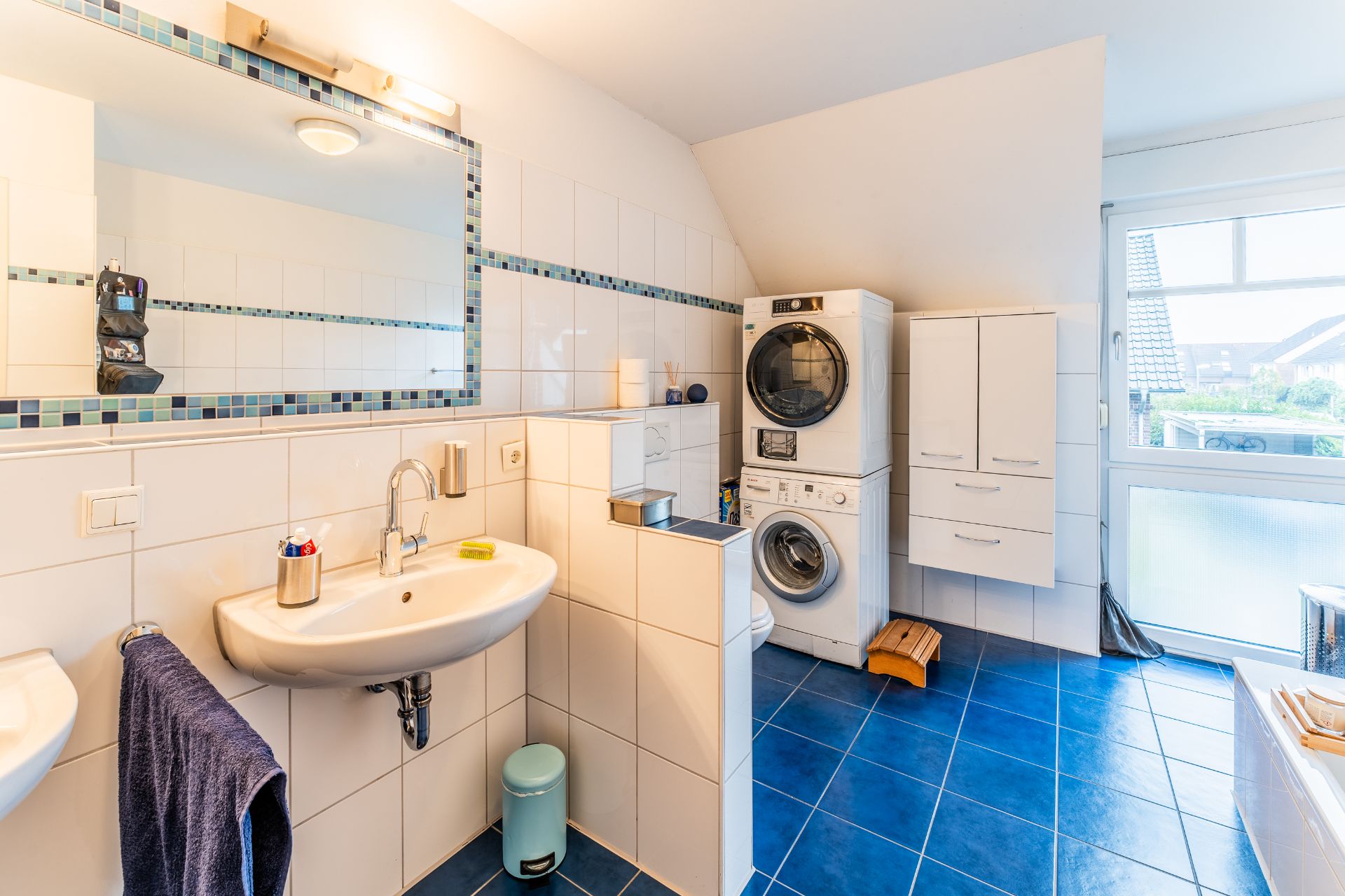 Modernes Badezimmer mit Waschbecken, Spiegel und Waschmaschine mit Trockner auf blau gefliestem Boden.