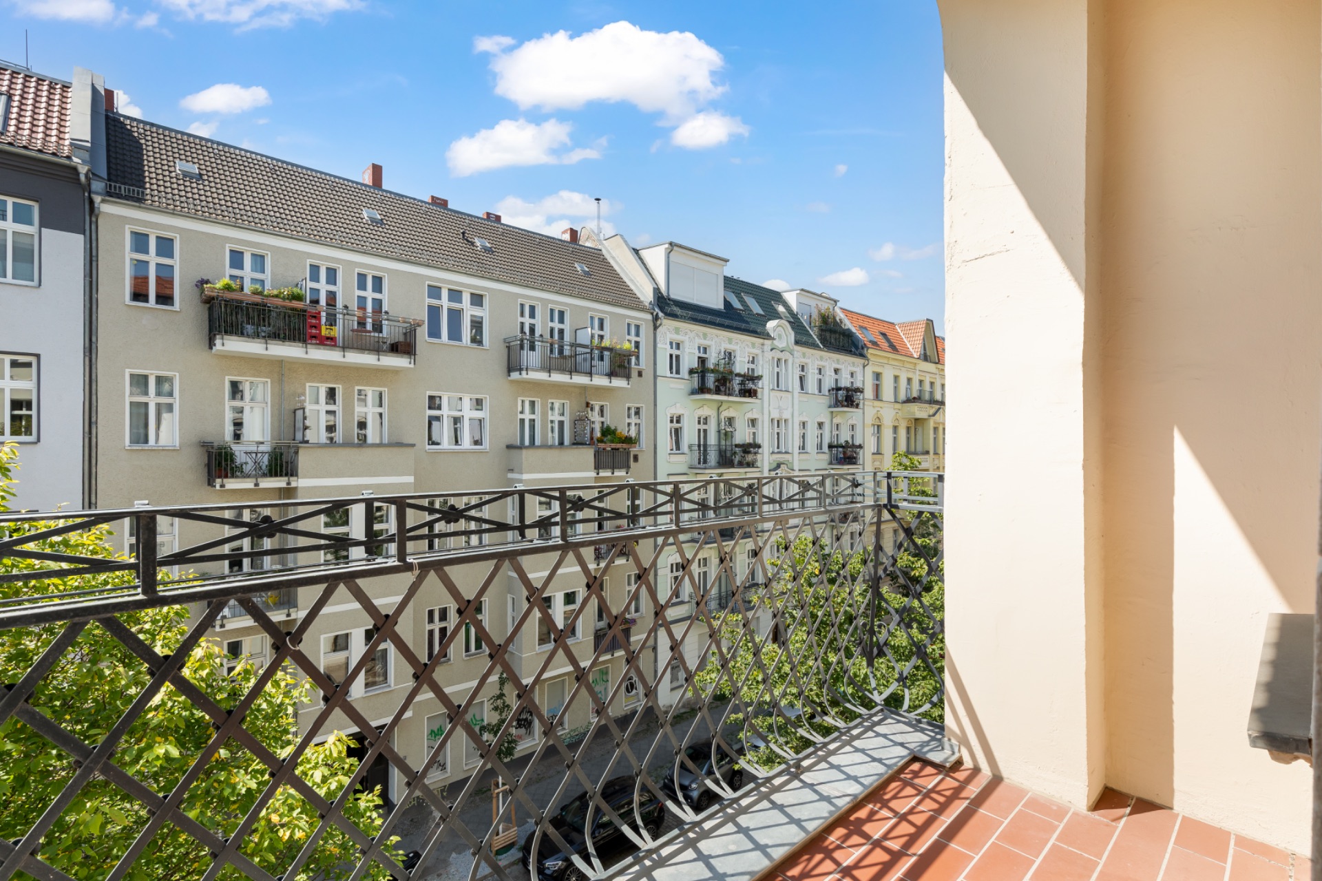 Blick von einem Balkon mit Gittergeländer auf gegenüberliegende Wohnhäuser und Bäume.