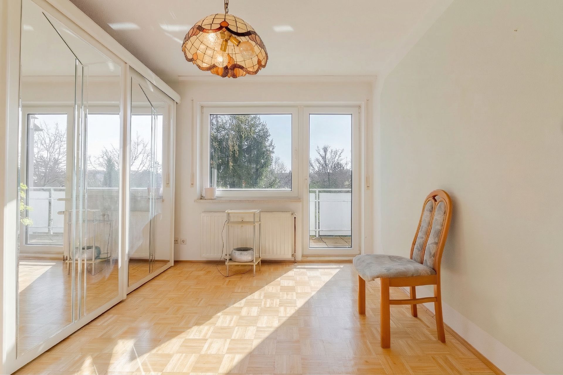 Heller Raum mit Parkettboden, Spiegelkleiderschrank, Stuhl und Fenstertür zum Balkon.