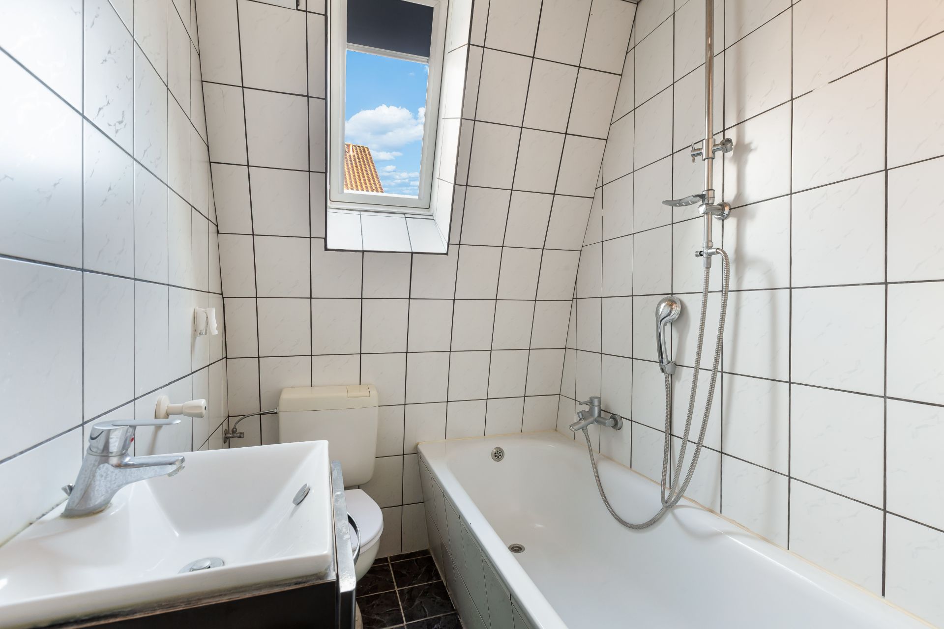 Kleines Badezimmer mit Badewanne, Waschbecken, Toilette und Fenster, weiße Fliesen an den Wänden.