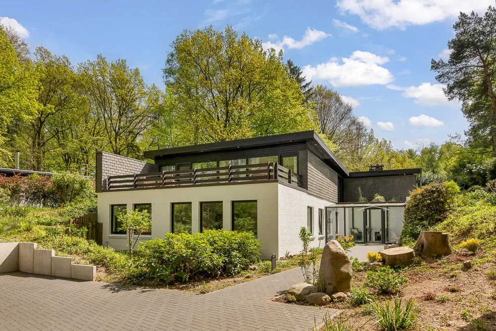 Modernes Wohnhaus mit Flachdach, großen Fenstern und Garten, umgeben von Bäumen.