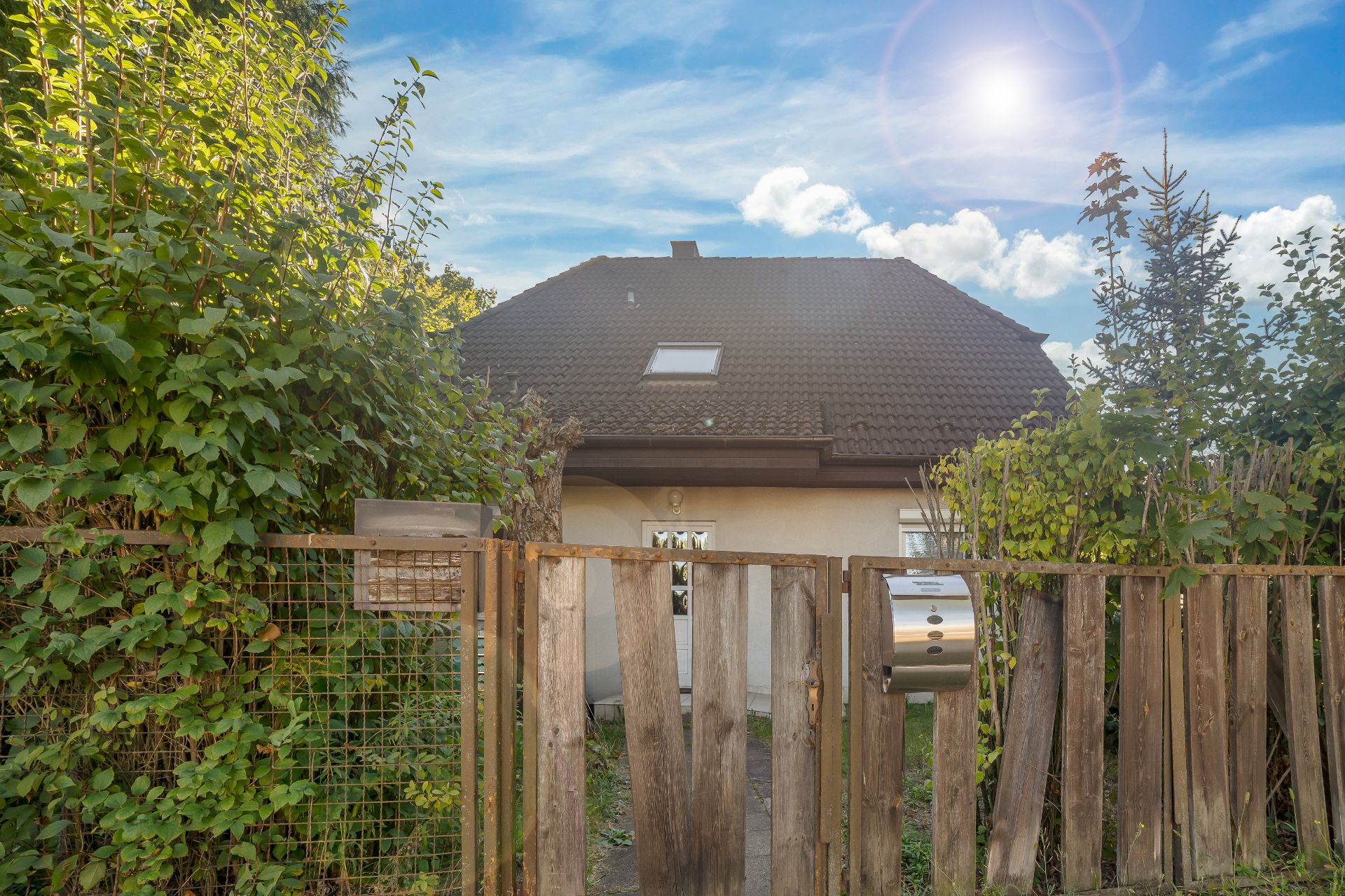 Haus mit braunem Dach hinter Holzzaun und viel Grün im Vordergrund.