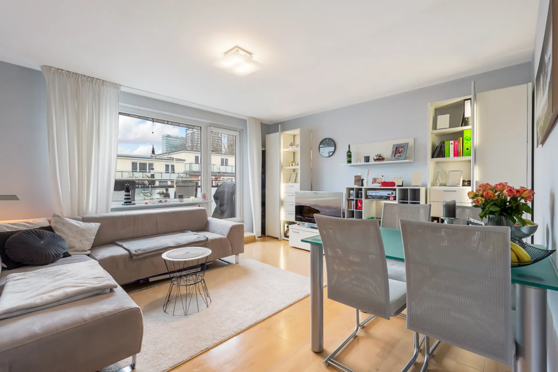 Helles Wohnzimmer mit Ecksofa, Esstisch, Regalen und großem Fenster.
