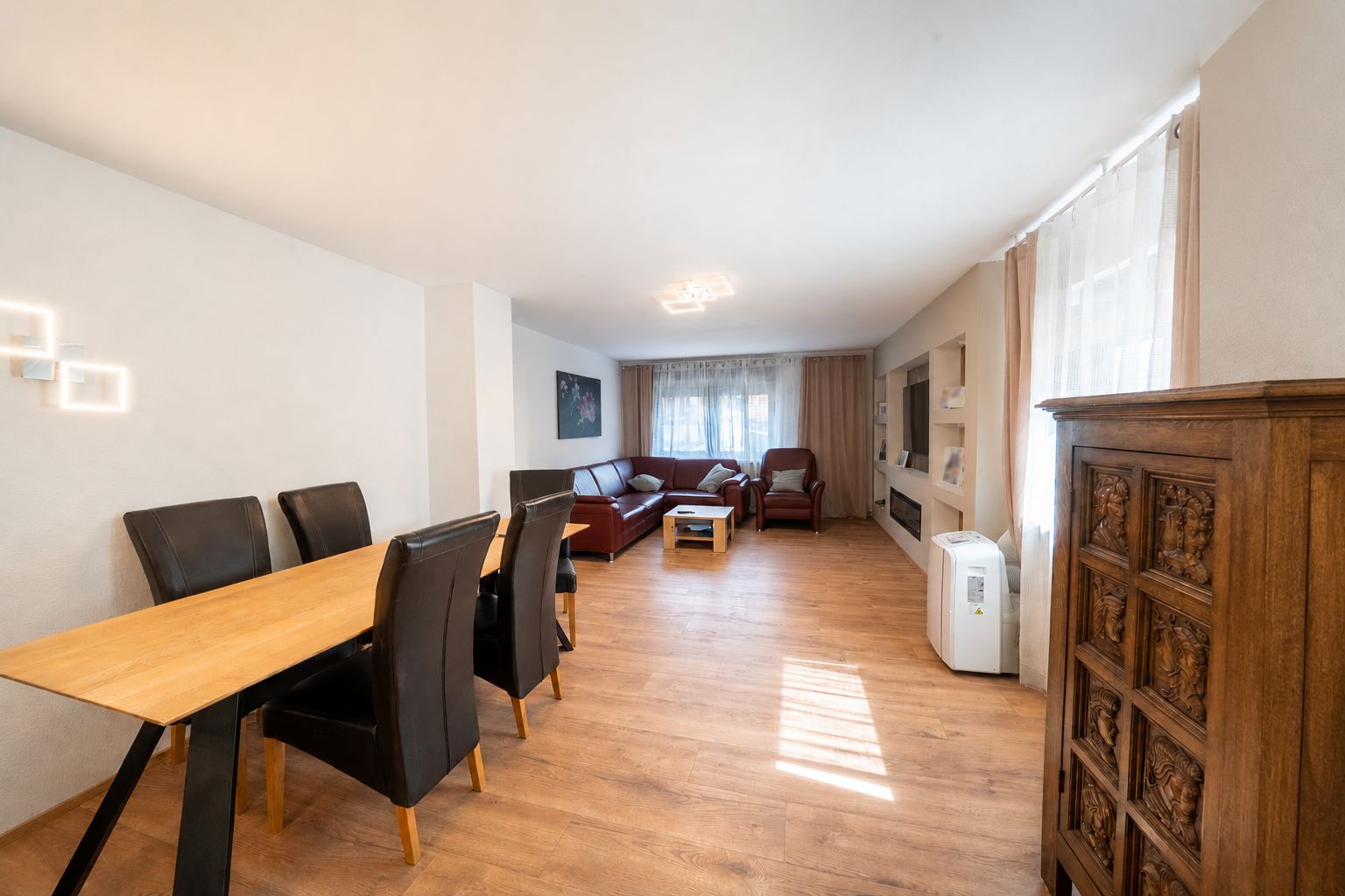 Wohnzimmer mit Esstisch, sechs Stühlen, roten Sofas und Fenster mit Vorhängen.