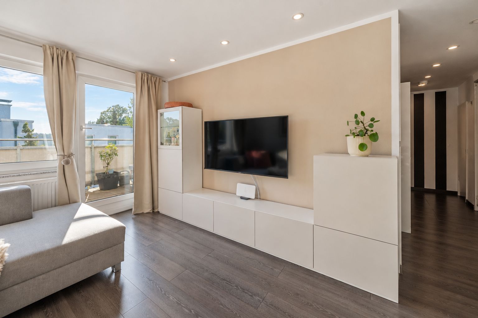 Helles Wohnzimmer mit Sofa, Fernseher an der Wand und Zugang zu einem Balkon.