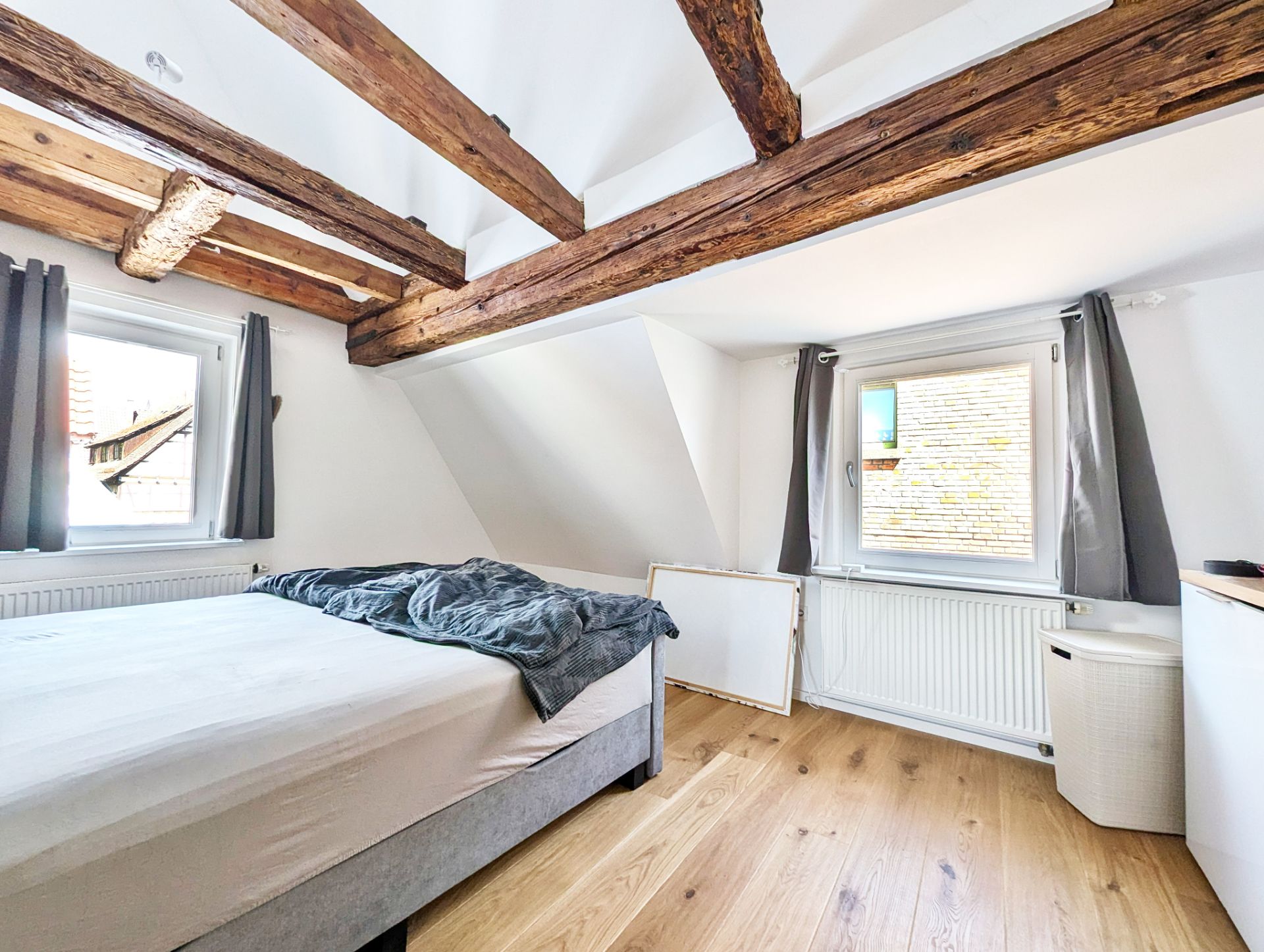 Helles Schlafzimmer mit Holzbalken, Bett, Holzboden und zwei Fenstern mit grauen Vorhängen.