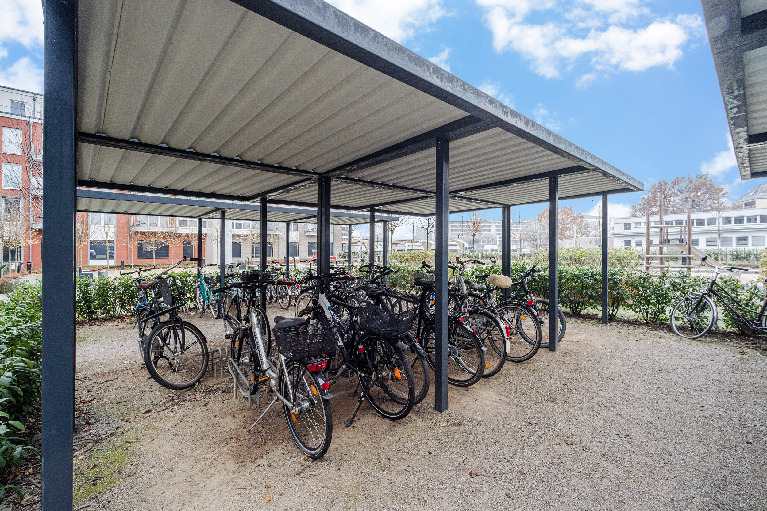 Überdachter Fahrradständer mit mehreren Fahrrädern auf einem Kiesplatz neben Wohnhäusern.