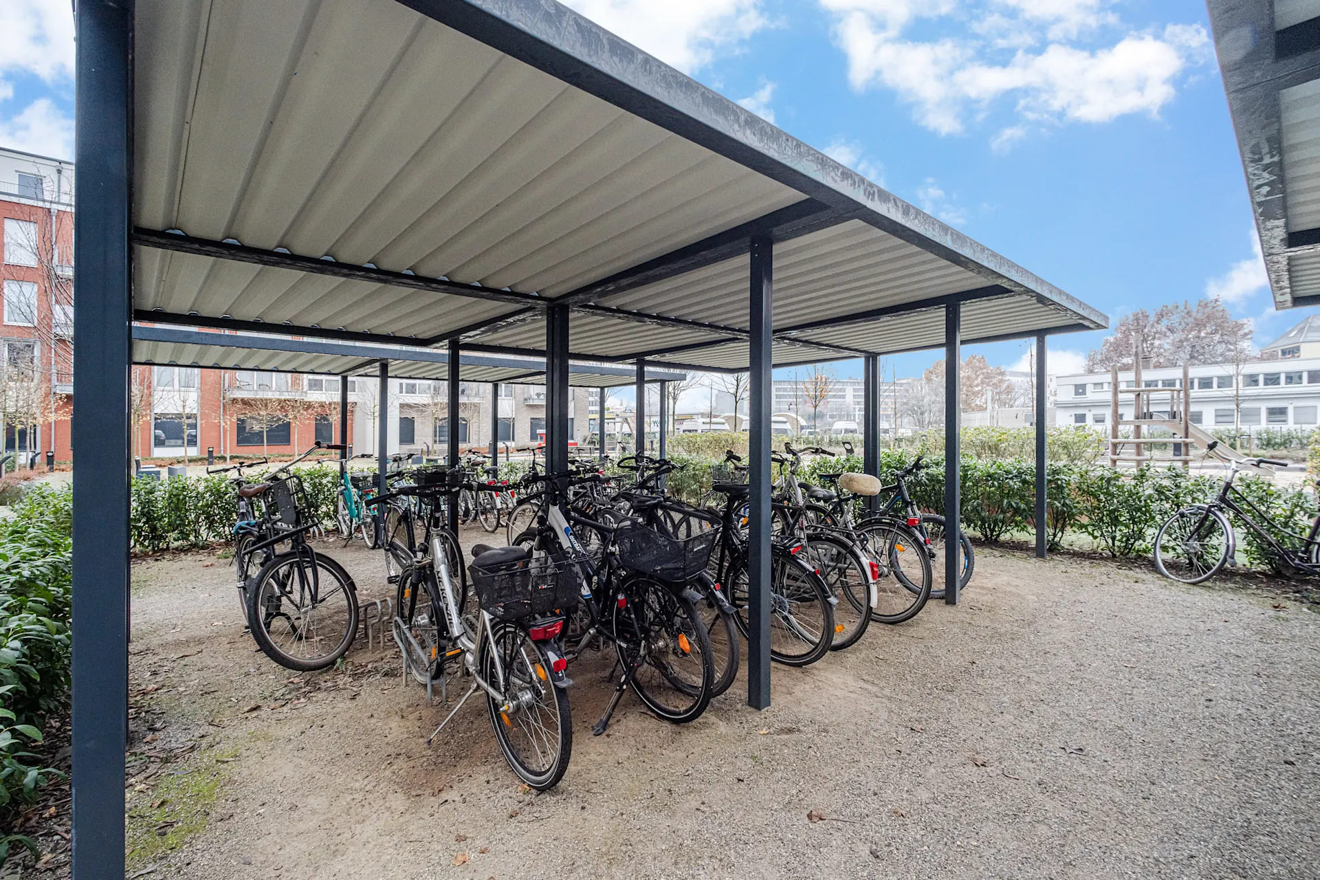 Überdachter Fahrradständer mit mehreren Fahrrädern auf einem Kiesplatz neben Wohnhäusern.