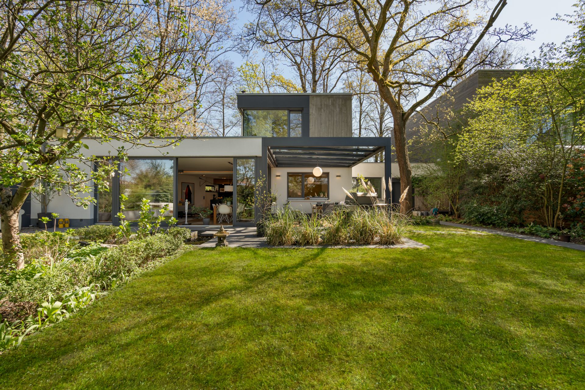 Modernes Wohnhaus mit großen Fenstern und Terrasse, umgeben von grünem Garten und Bäumen.