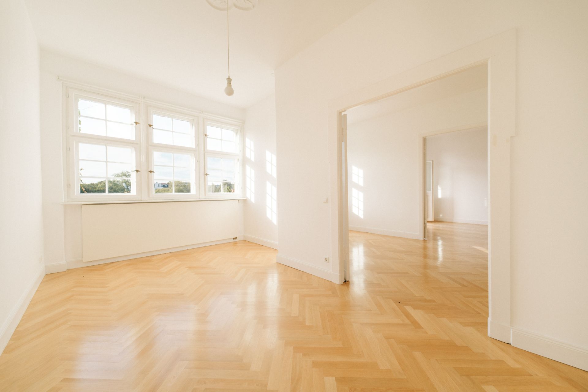 Heller Raum mit großen Fenstern, Parkettboden und offener Verbindung zu einem weiteren Zimmer.
