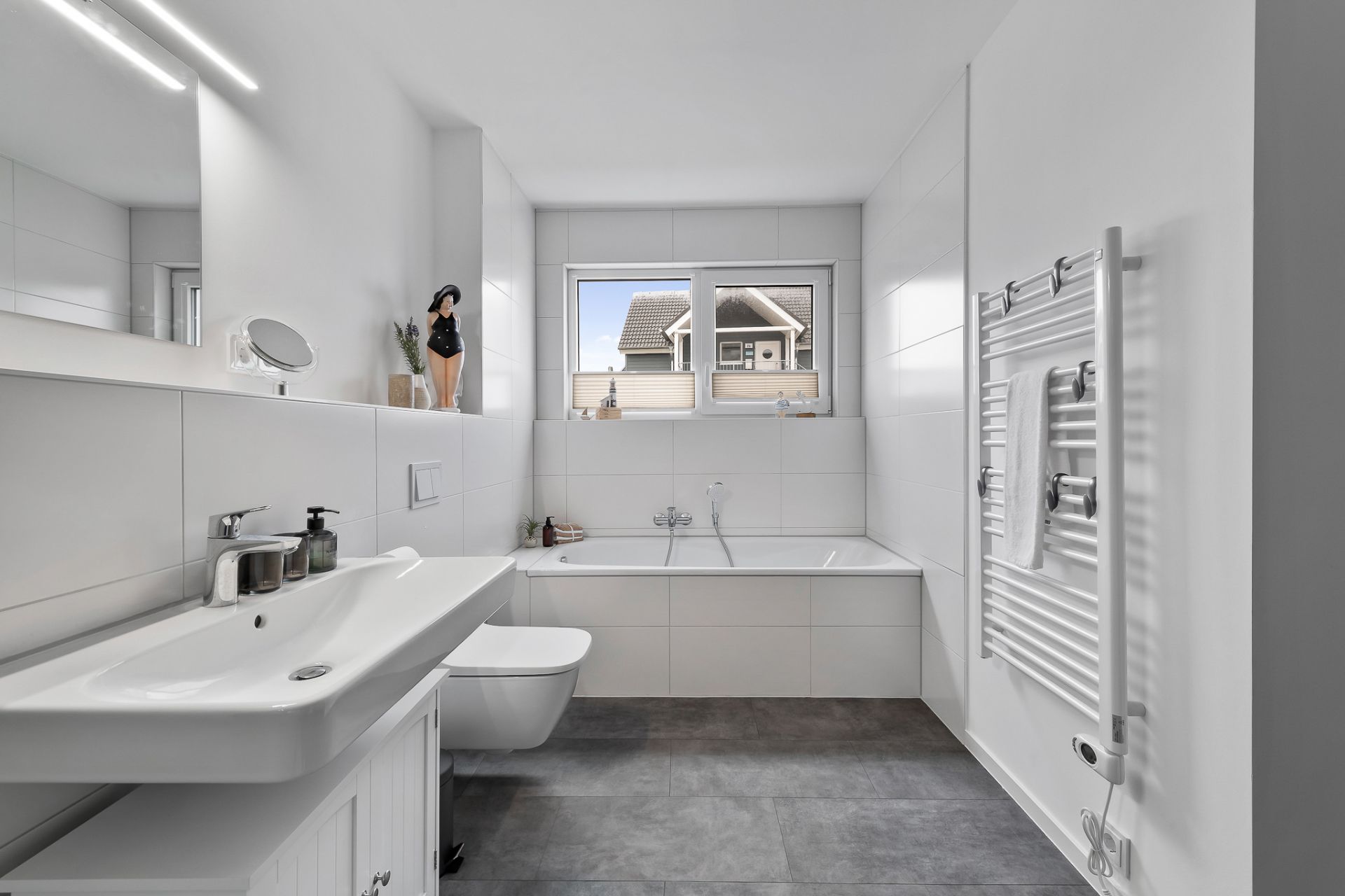 Modernes Badezimmer mit Badewanne, Waschbecken, WC und Fenster, Handtuchheizkörper an der Wand.