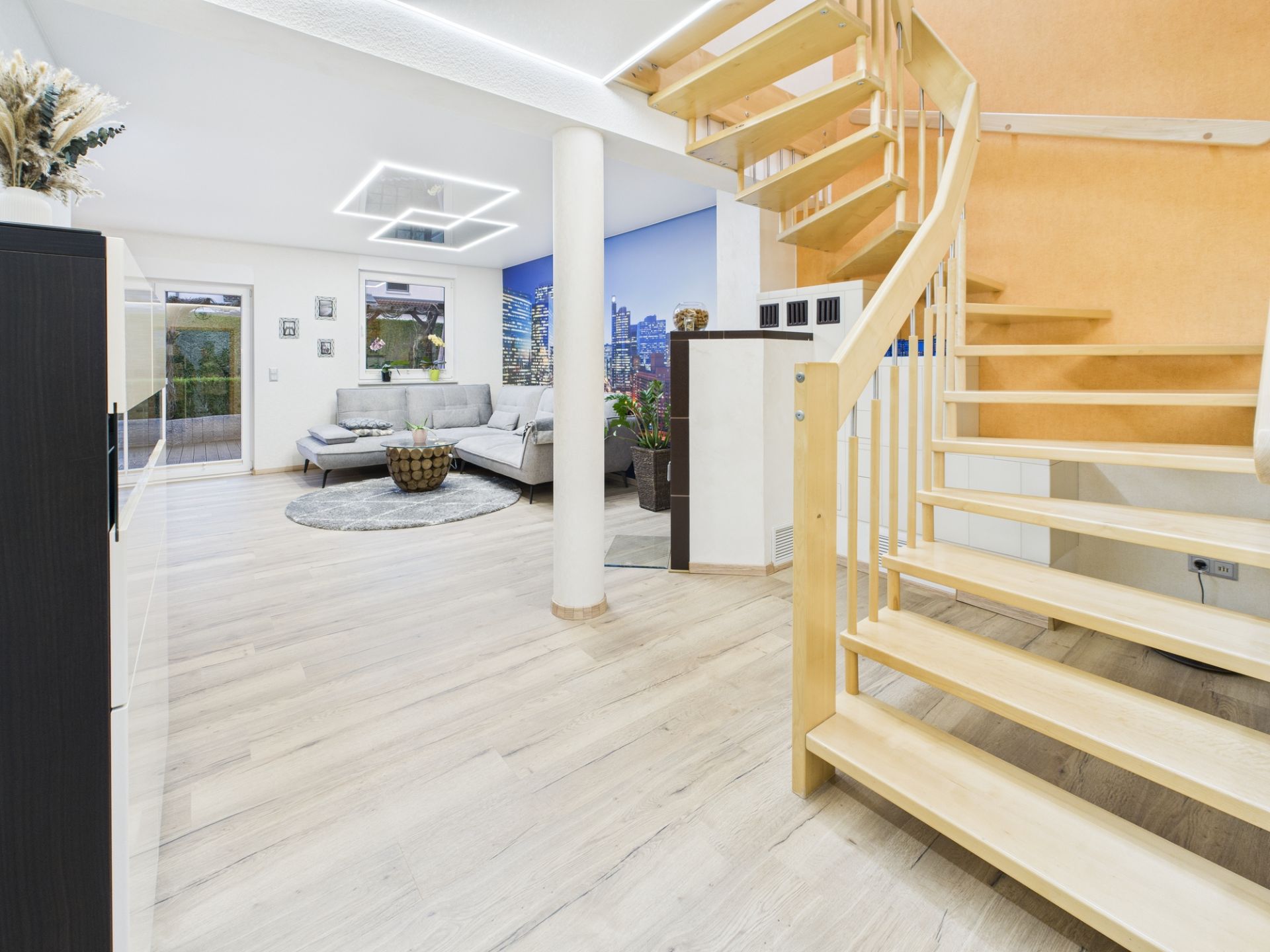 Helles Wohnzimmer mit Holztreppe, grauem Sofa, Couchtisch und großem Wandbild.