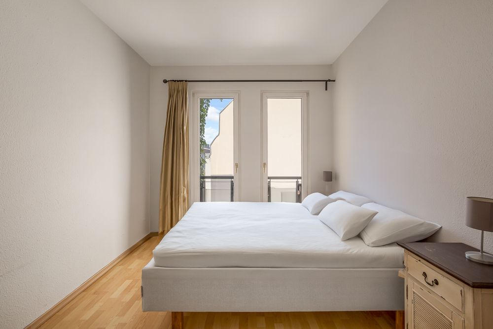 Helles Schlafzimmer mit Doppelbett, Nachttisch und Zugang zu einem kleinen Balkon.