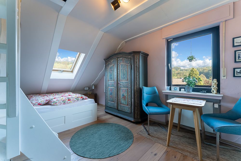 Gemütliches Schlafzimmer mit Bett, Schrank, zwei blauen Stühlen und Tisch, Fenster mit Blick ins Grüne.