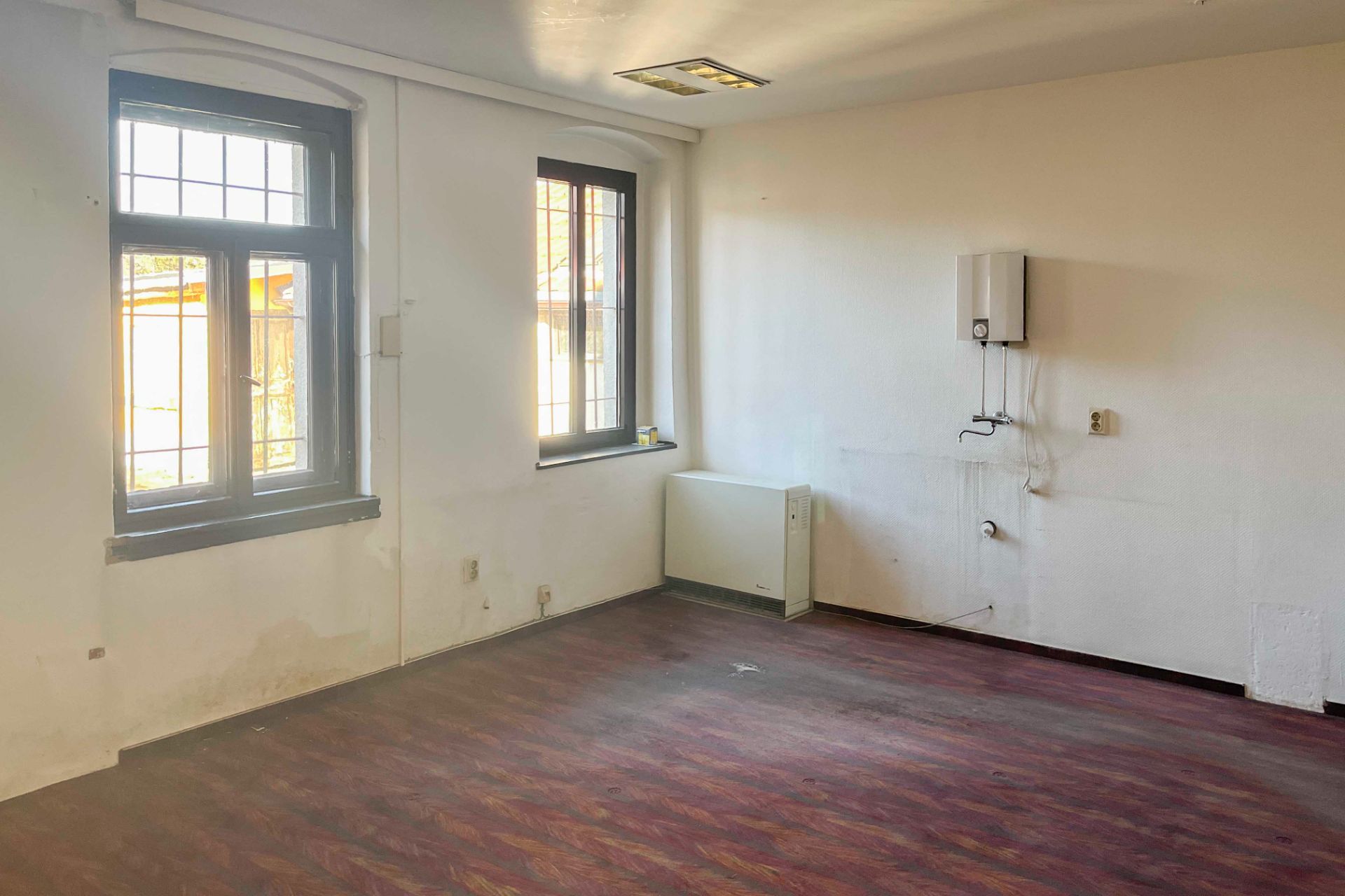 Leeres Zimmer mit zwei Fenstern, Heizkörper und Wandgerät, dunkler Boden.