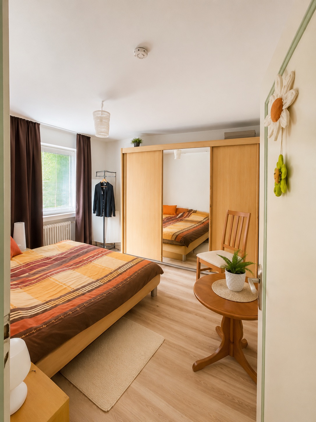Helles Schlafzimmer mit Doppelbett, Kleiderschrank mit Spiegel und kleinem Tisch mit Pflanze.