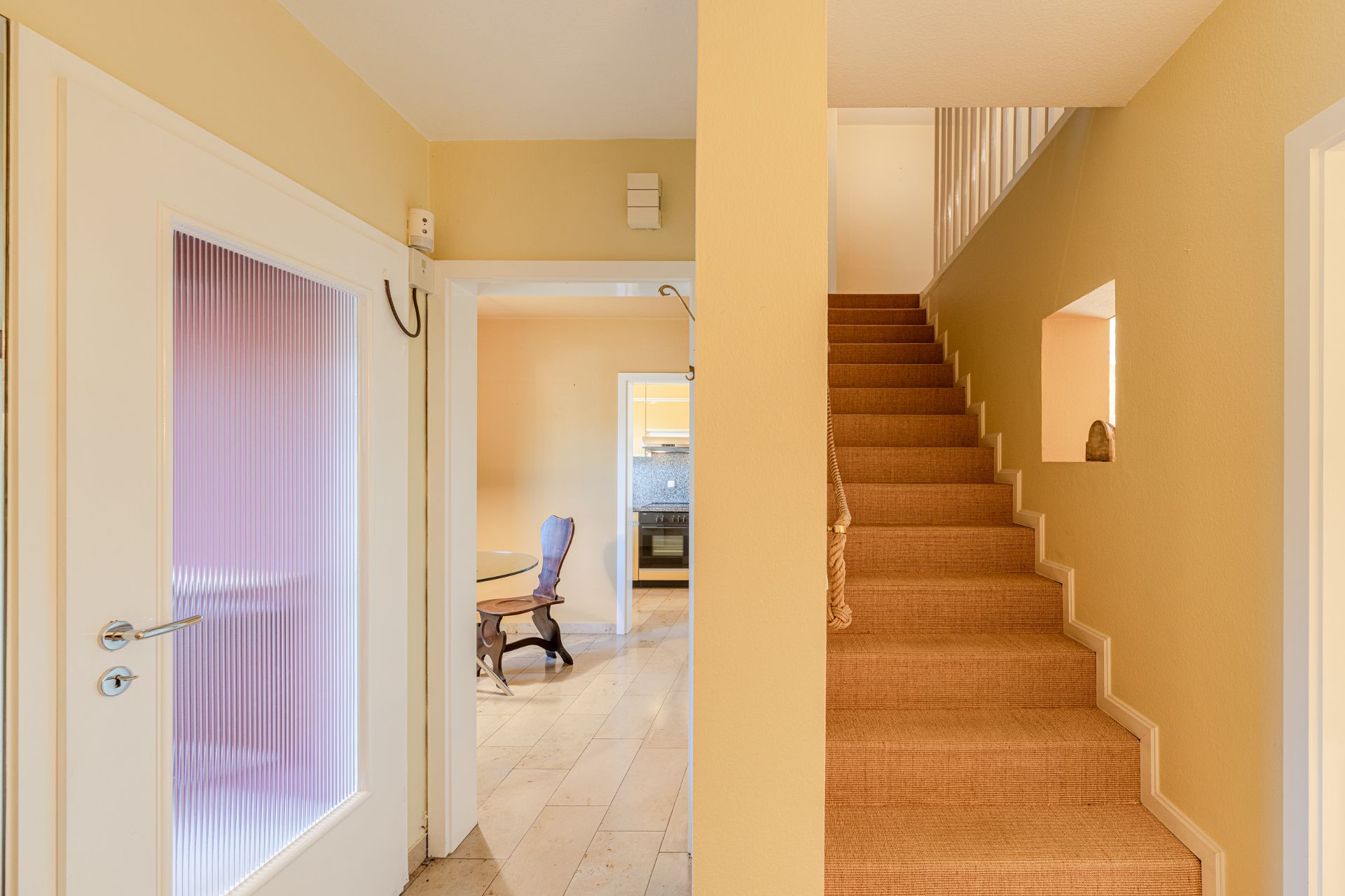 Flur mit Treppe nach oben, Tür mit geriffeltem Glas und Blick ins Esszimmer mit Stuhl und Tisch.