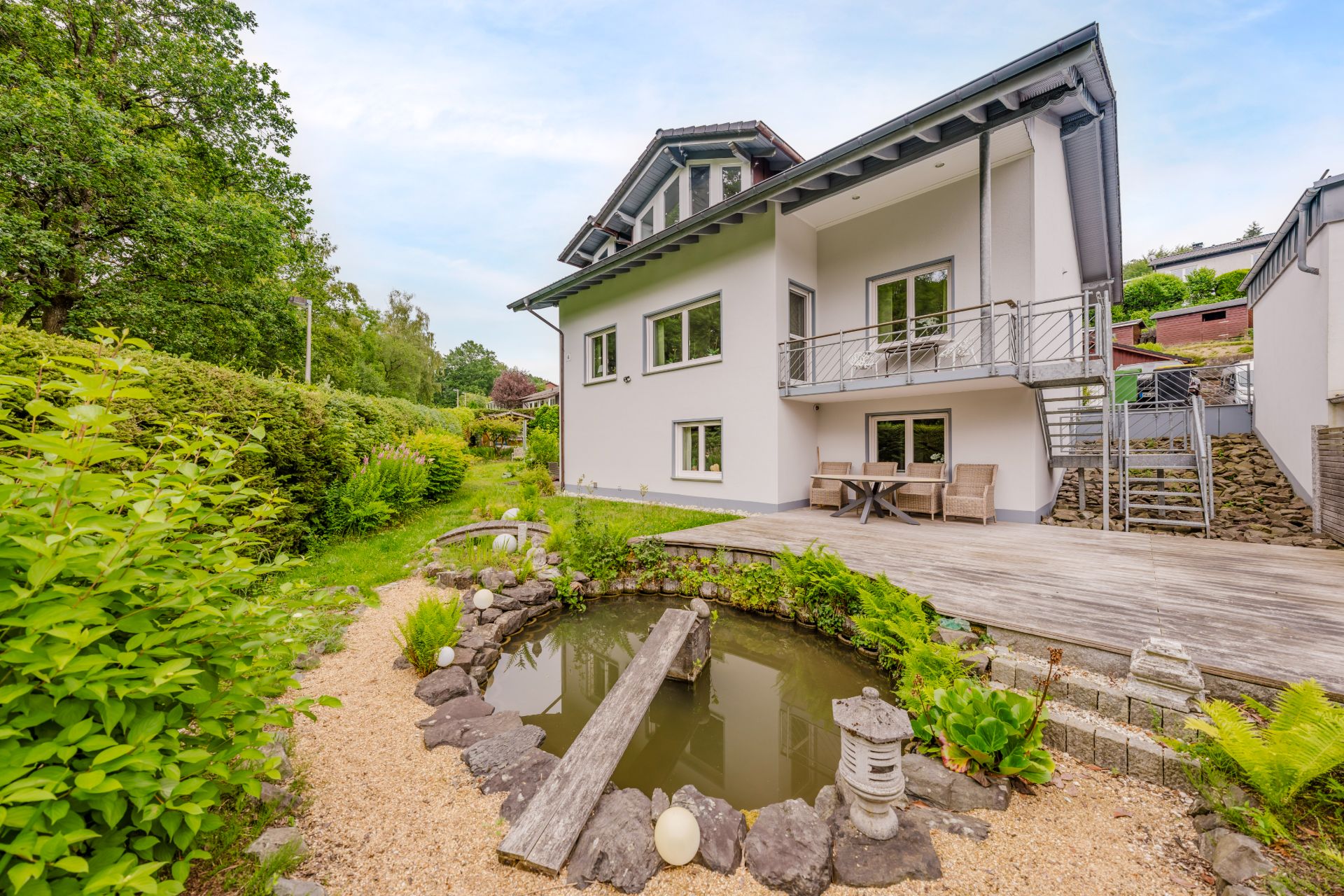Modernes Wohnhaus mit Holzbalkon, Terrasse und kleinem Gartenteich im gepflegten Garten.