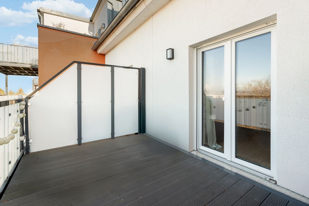 Balkon mit Glasschutzwand und Doppeltür, Blick auf Hausfassade und blauen Himmel.