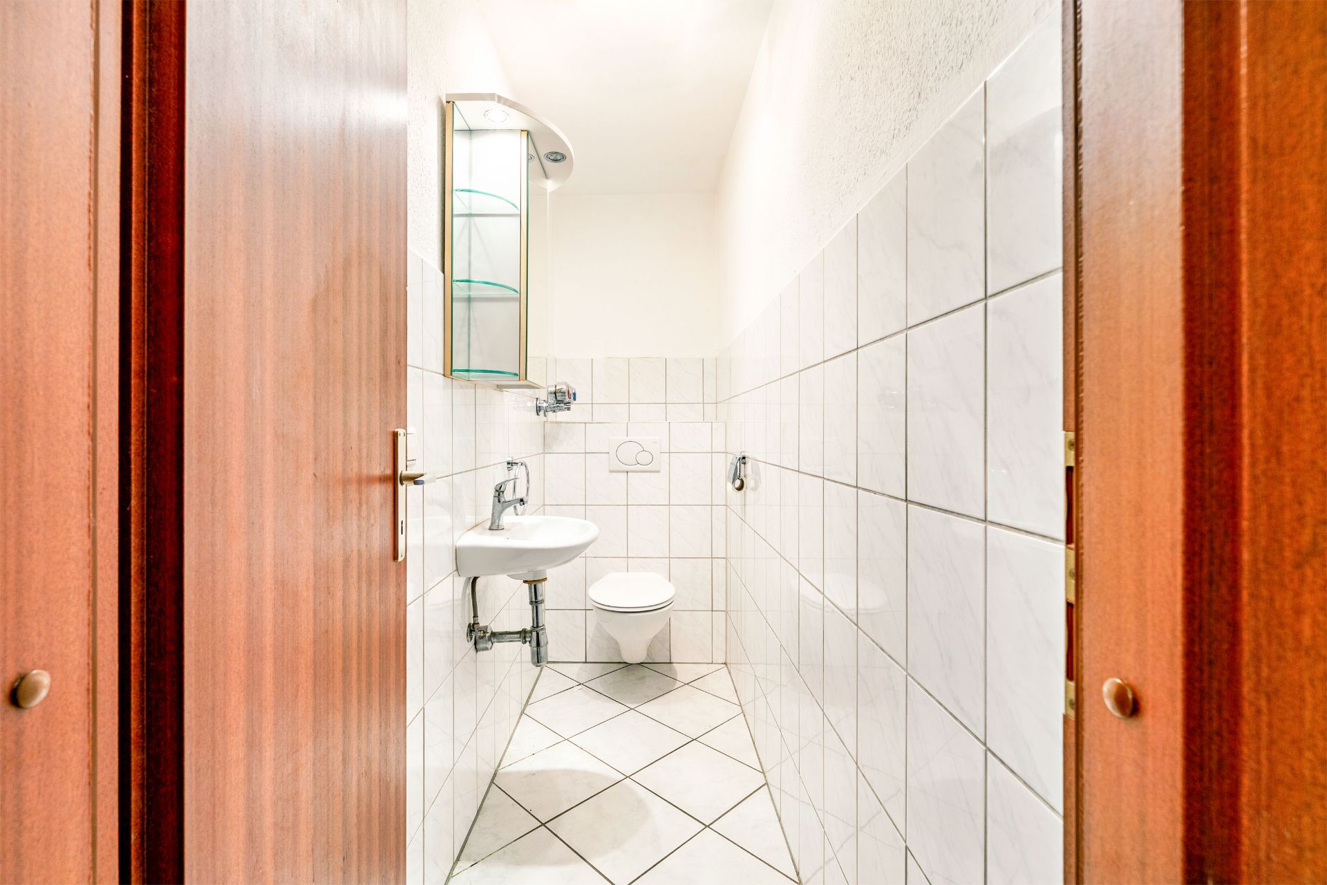 Gefliestes kleines Badezimmer mit Wandwaschbecken, Spiegelregal und Wandtoilette.