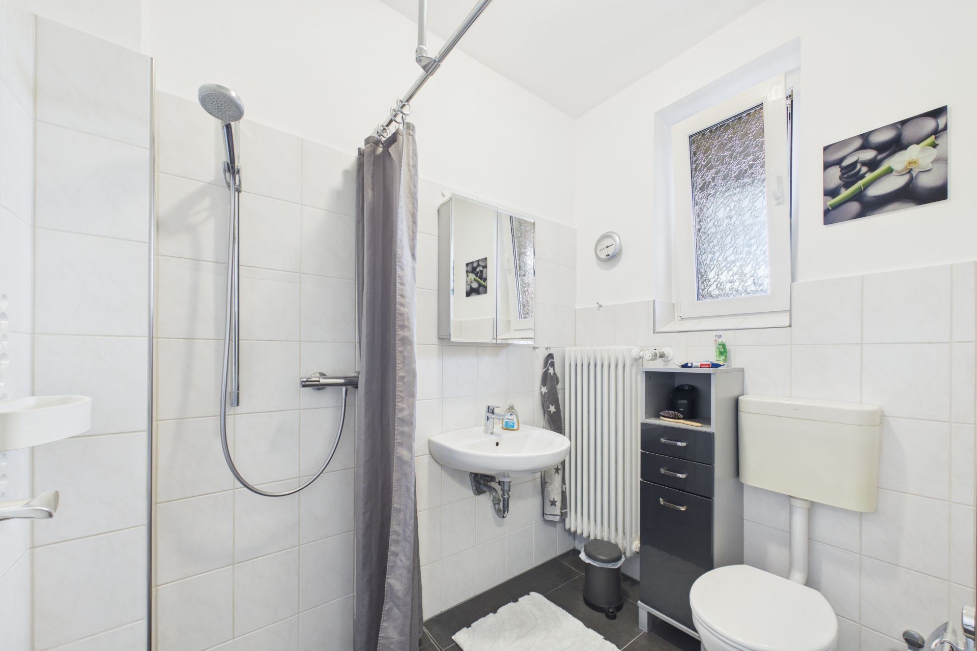 Helles Badezimmer mit Dusche, Waschbecken, Toilette, Fenster und kleinem Schrank.
